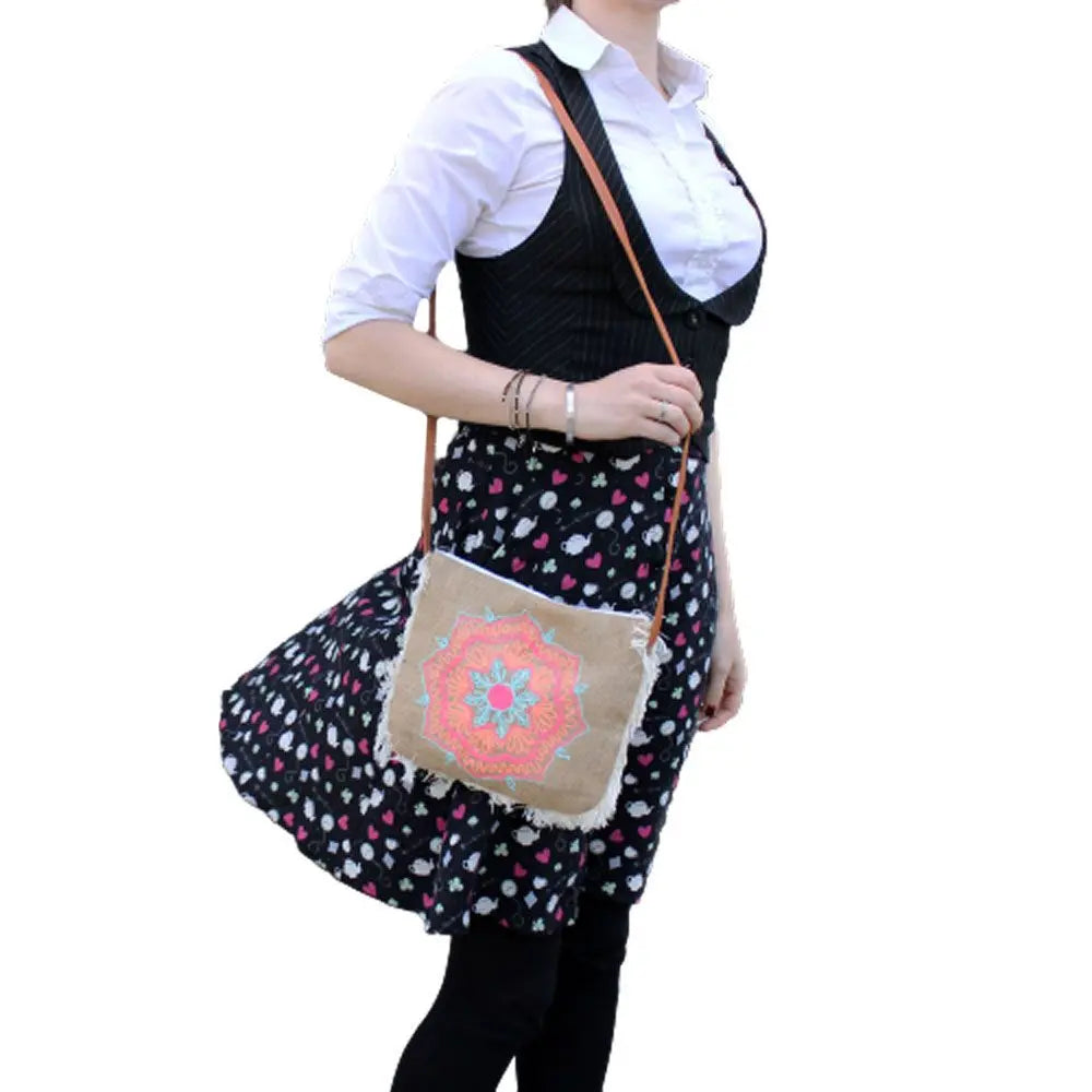 Bolso bandolera de arpillera con flecos y bordado de mandala en tonos rosa y azul, llevado por una persona.