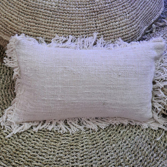 Funda de cojín rectangular de lino color beige con flecos decorativos en los bordes sobre superficie de fibra natural