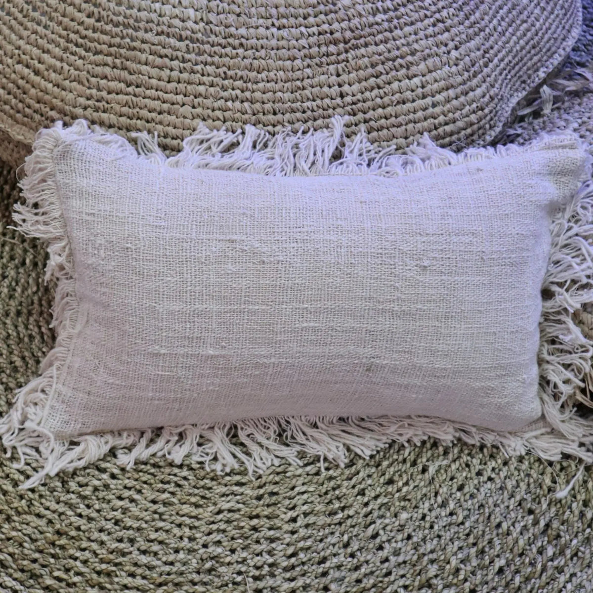 Funda de cojín rectangular de lino color beige con flecos decorativos en los bordes sobre superficie de fibra natural