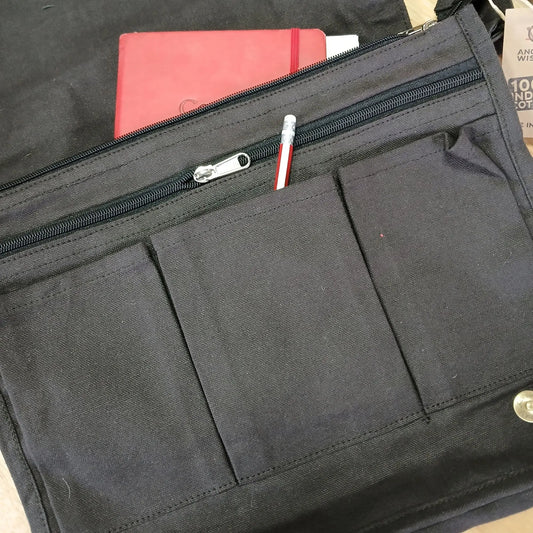 Bolsa mensajera de lona gris carbón con bolsillos frontales, cremallera, un lápiz y una libreta roja en su interior.