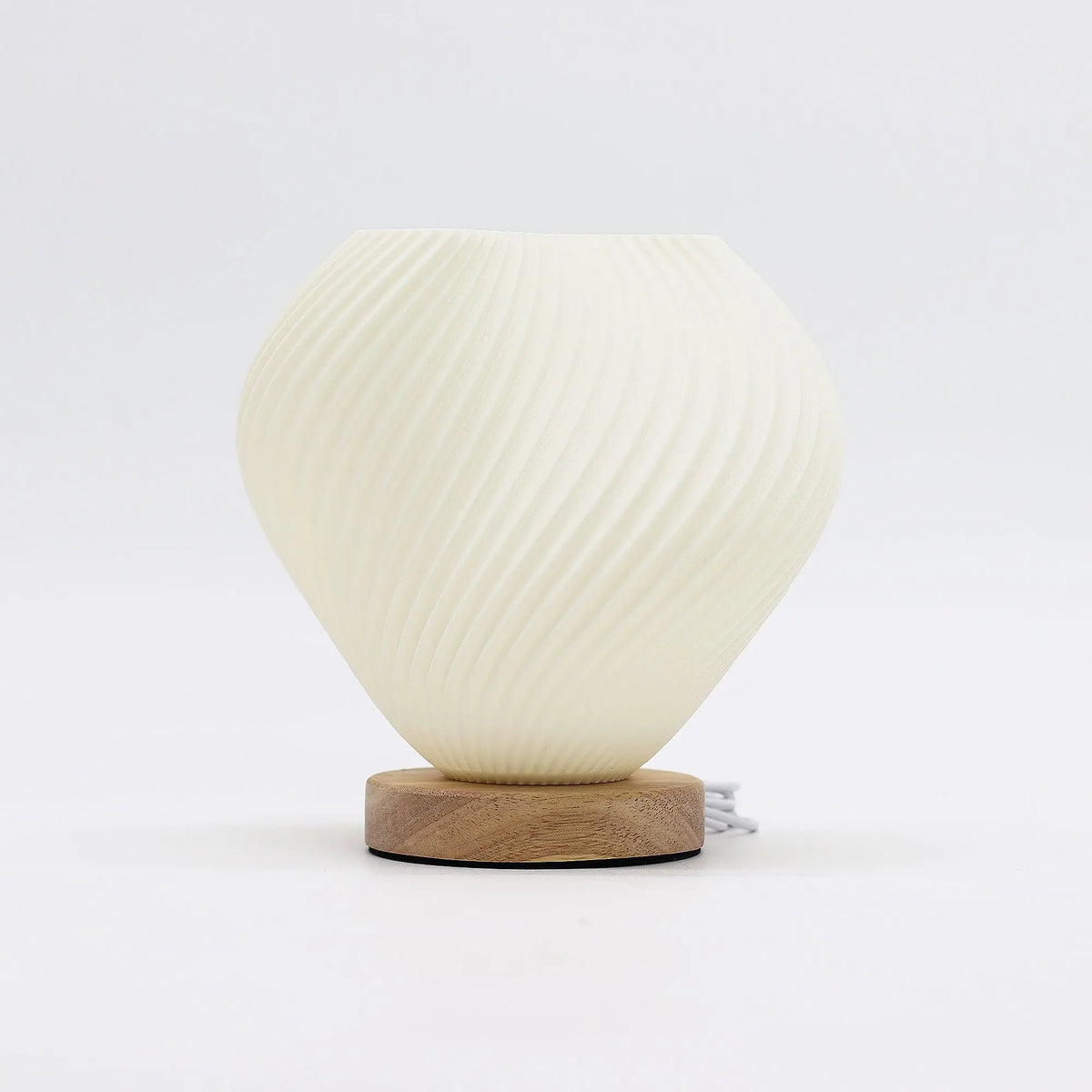Lámpara de mesa en forma de concha espiral color crema sobre base circular de madera clara, con cable USB visible.