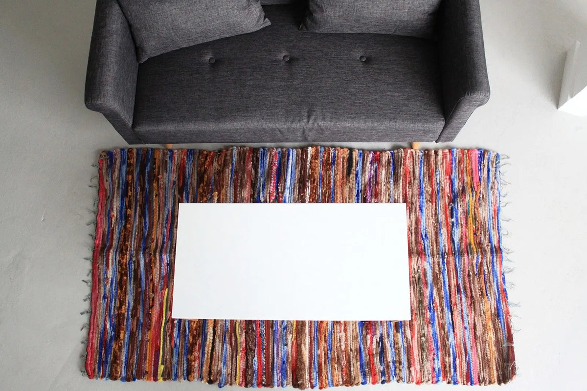 Alfombra de trapo rectangular (105x185cm) con rayas verticales multicolor y texturas artesanales bajo una mesa blanca.