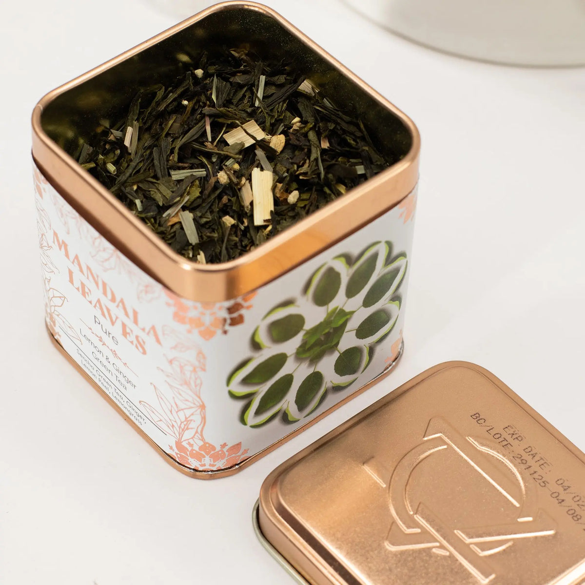 Lata cuadrada color cobre con té verde, trozos de jengibre y limón, junto a su tapa metálica sobre fondo blanco.