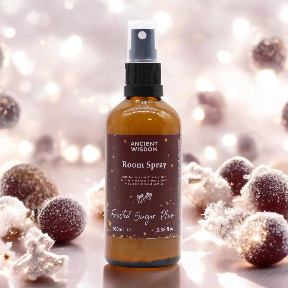 Spray de fragancia de 100 ml con etiqueta marrón, rodeado de ciruelas escarchadas y luces navideñas desenfocadas.