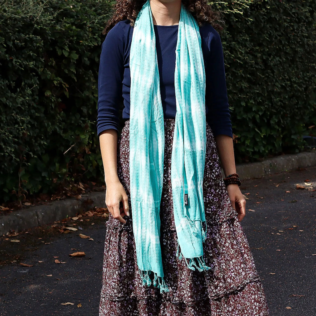 Estola larga color turquesa con rayas blancas, estilo boho, tejido ligero con flecos en los extremos sobre falda floral.
