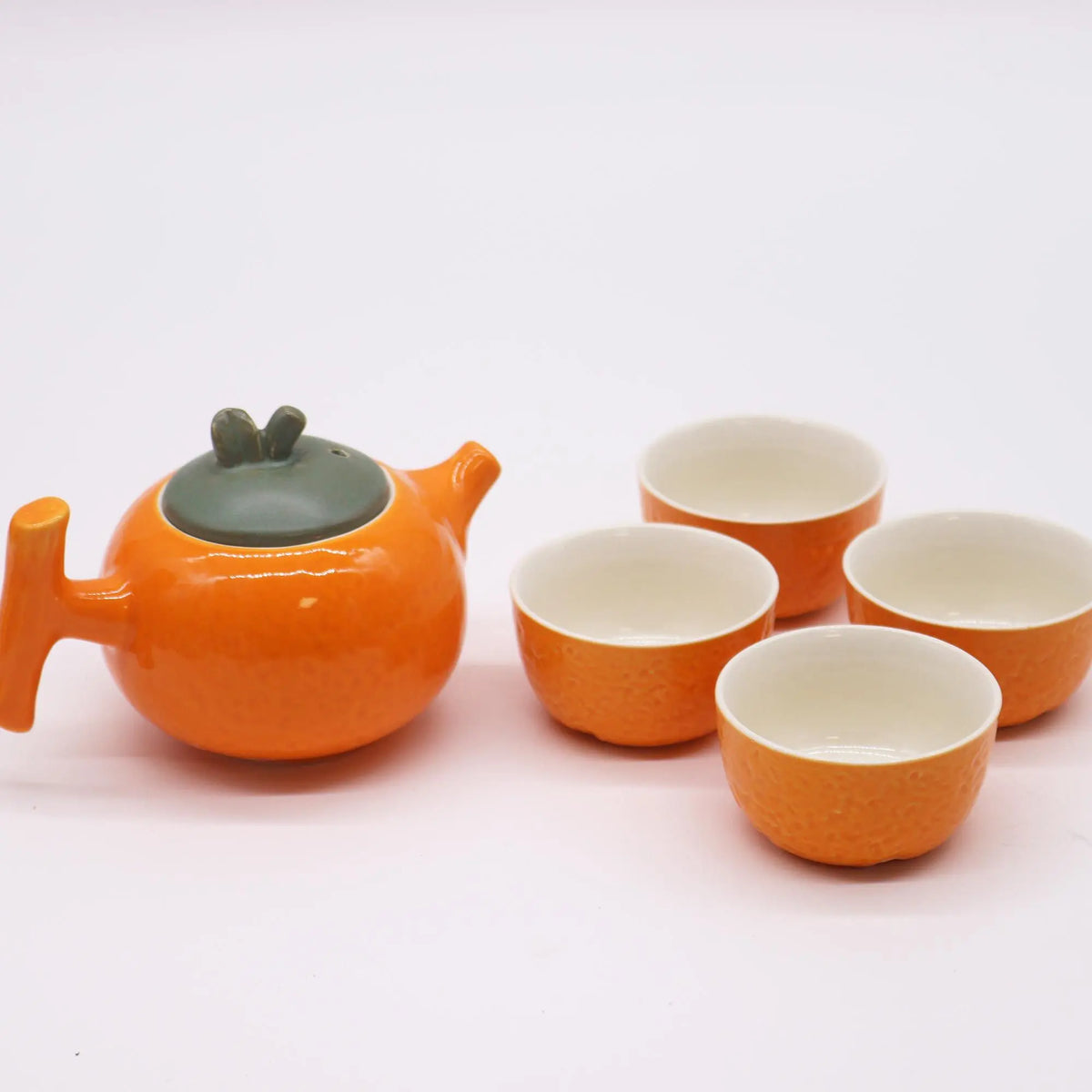 Juego de té de cerámica naranja con textura, tetera con tapa verde y cuatro tazas pequeñas sobre fondo blanco.