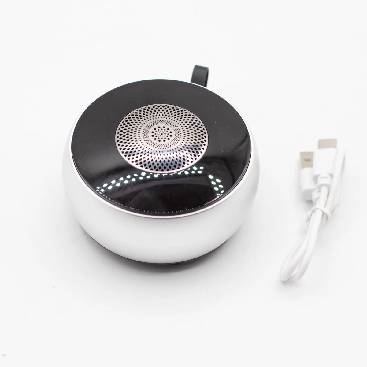 Altavoz Bluetooth mini, plateado y negro, forma circular, rejilla con diseño mandala y cable de carga USB incluido.