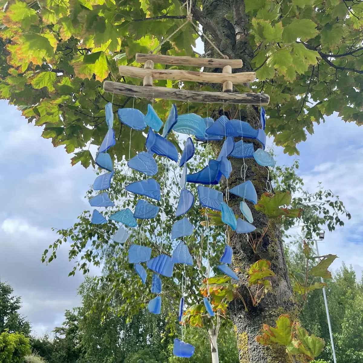 Carrillón de piezas de vidrio reciclado azul colgando de una estructura de madera rústica en un árbol.
