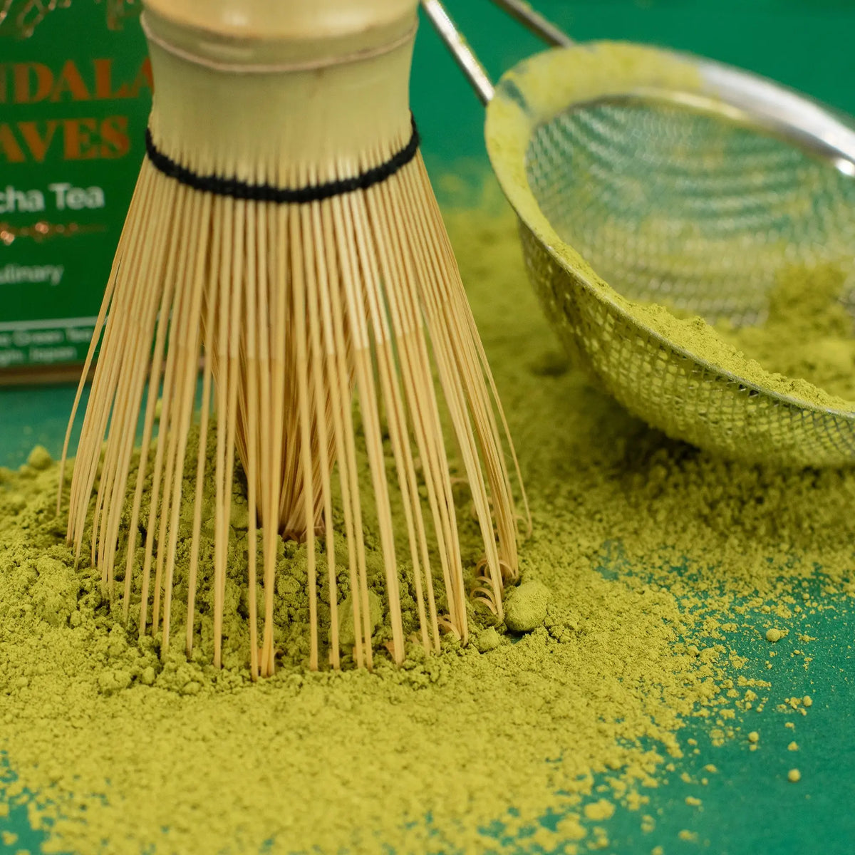 Batidor de bambú sobre polvo de té matcha verde con un colador metálico sobre un fondo verde.