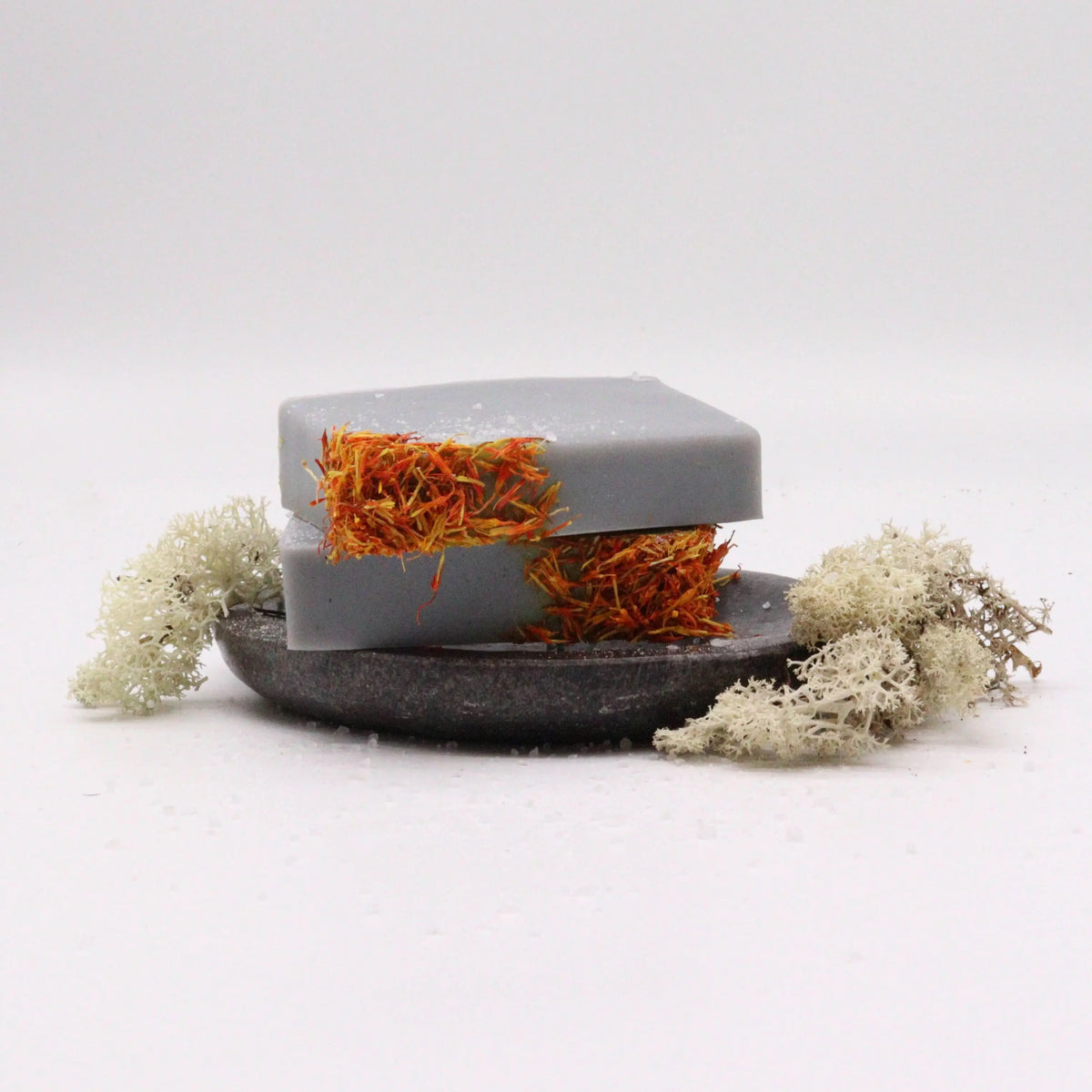 Dos barras de jabón gris rectangulares con pétalos naranjas, sobre un plato oscuro junto a líquenes blancos.