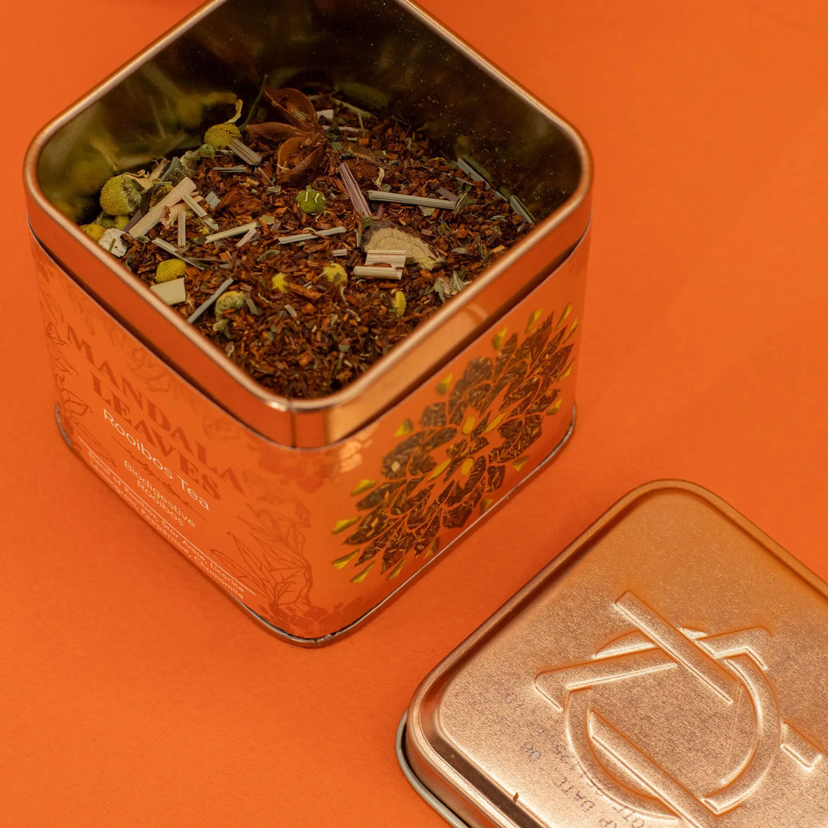 Lata metálica cuadrada abierta con mezcla de té rooibos hierbas y flores sobre fondo naranja de estilo artesanal