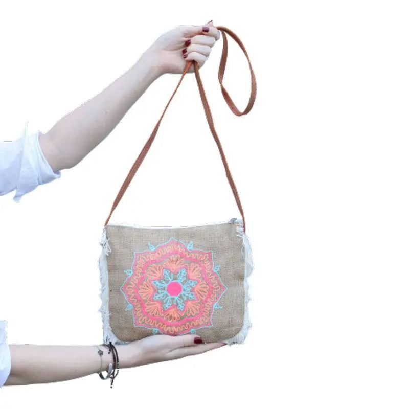 Bolso de arpillera con flecos laterales, bordado de mandala en tonos rosa y azul, y correa larga de cuero marrón.
