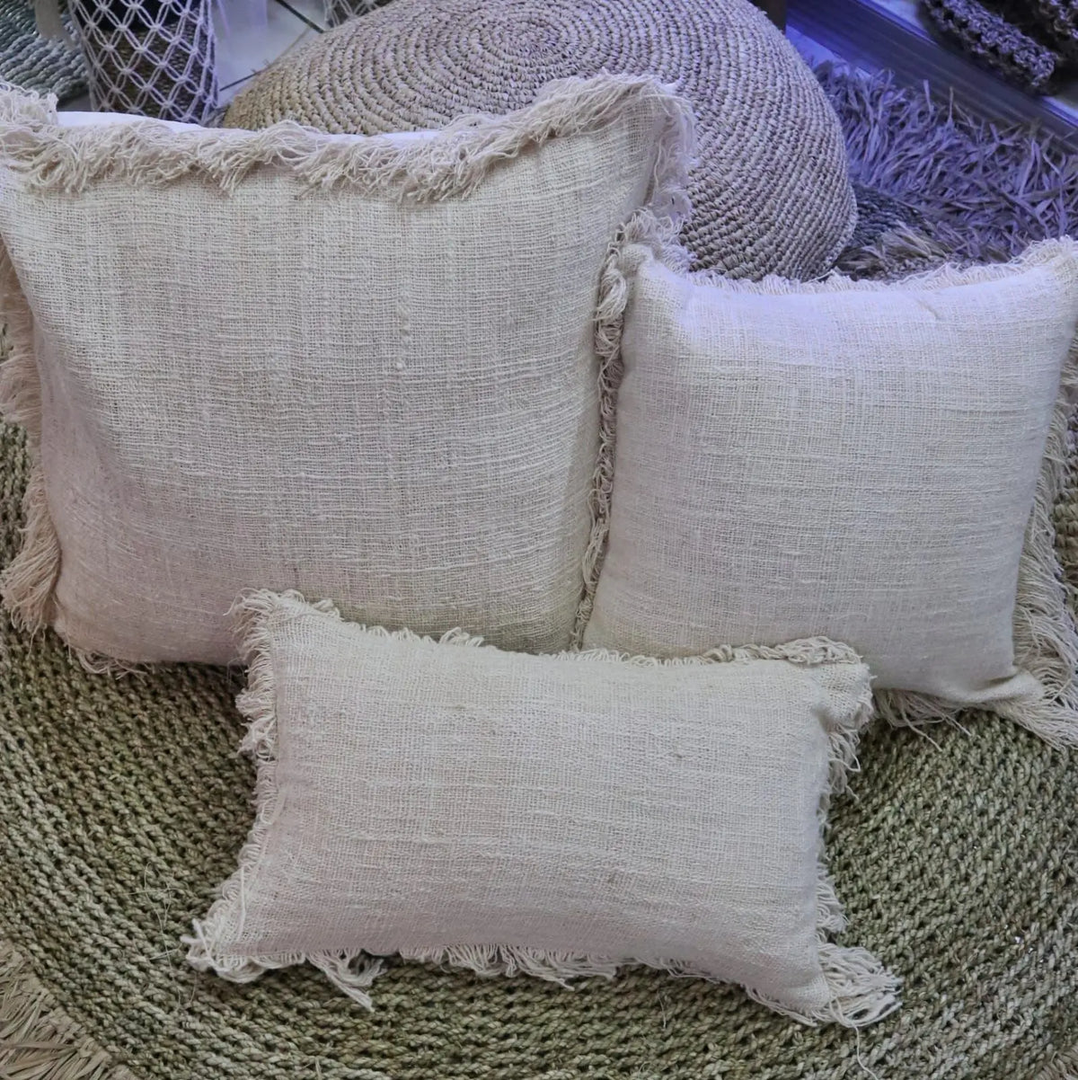 Tres fundas de cojín de lino color beige con textura rústica y flecos en los bordes sobre una alfombra de yute.