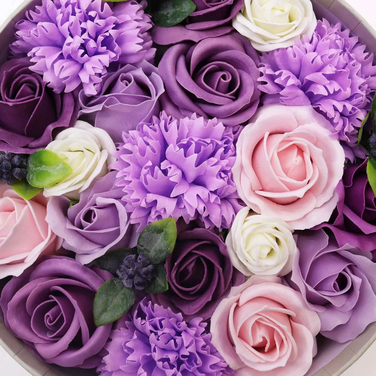 Caja redonda llena de rosas y claveles de jabón en tonos morados, lilas y rosas con pequeñas hojas verdes.