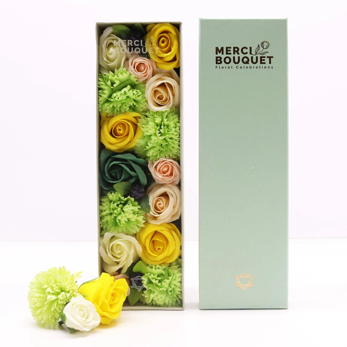 Caja rectangular abierta con flores de jabón amarillas, verdes y crema. Tapa verde a un lado sobre fondo blanco.