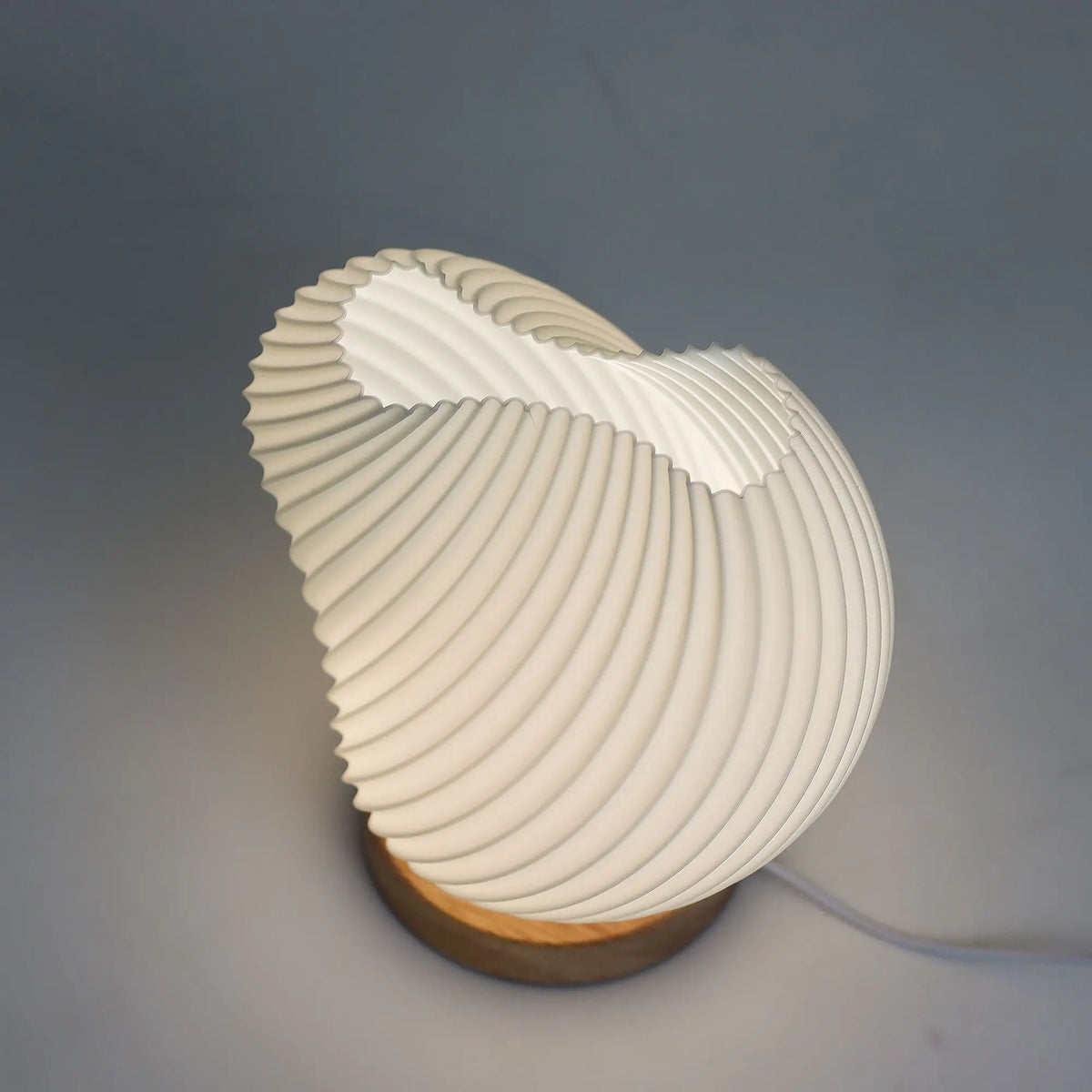 Lámpara blanca de sobremesa con forma de concha espiralada, pantalla estriada sobre base circular de madera clara.