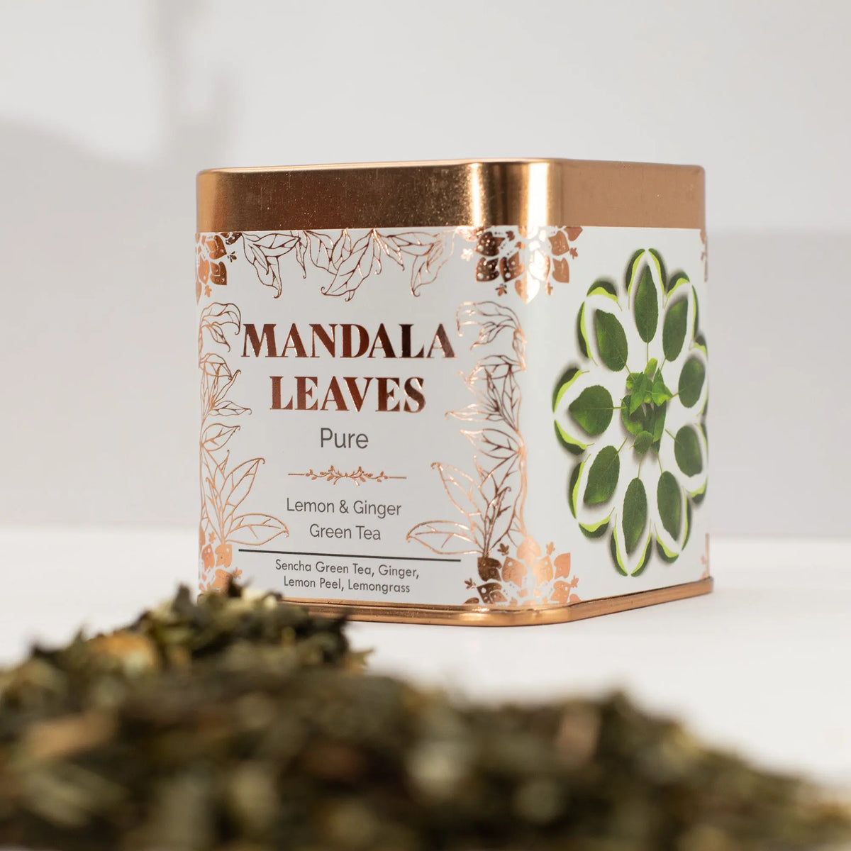 Lata cuadrada blanca y cobre de té verde con limón y jengibre, junto a hojas de té seco sobre superficie blanca.