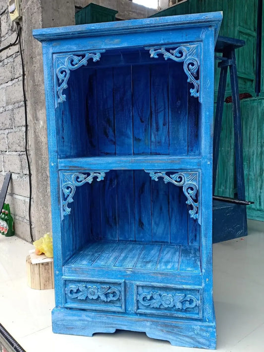 Mueble de madera azul con estantes abiertos, detalles tallados en estilo rústico y dos cajones inferiores decorativos.