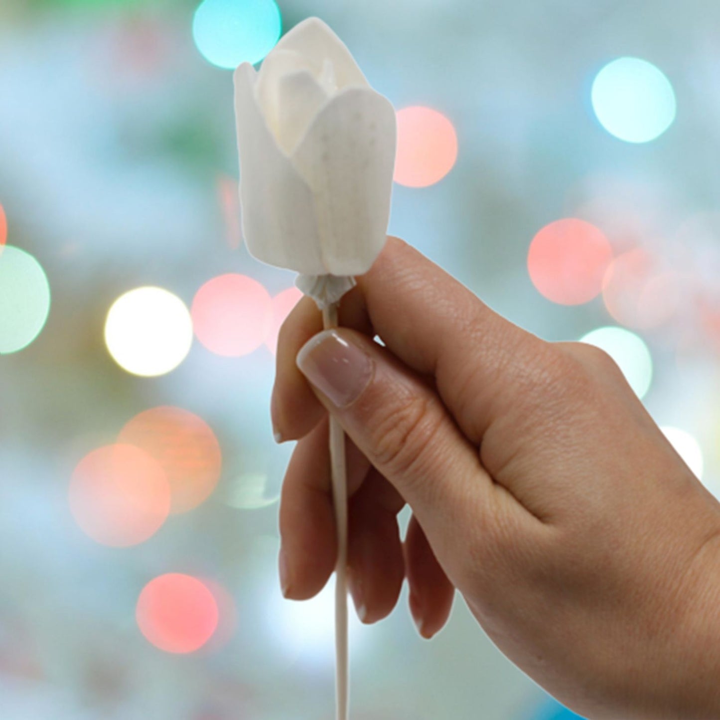 Mano sosteniendo un difusor artesanal con forma de tulipán blanco de madera de sola y tallo de caña. Fondo con luces.