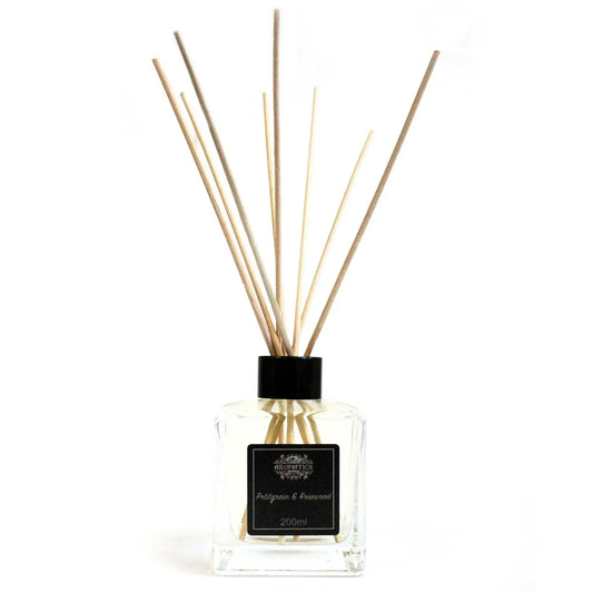 Difusor de aceites en frasco de vidrio cuadrado transparente, tapa negra y varillas de madera sobre fondo blanco.