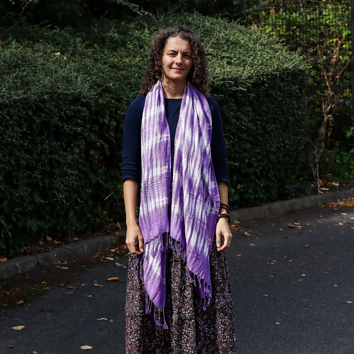 Mujer con estola larga lavanda y blanca de estilo boho, con diseño de rayas y flecos, sobre fondo de jardín exterior.