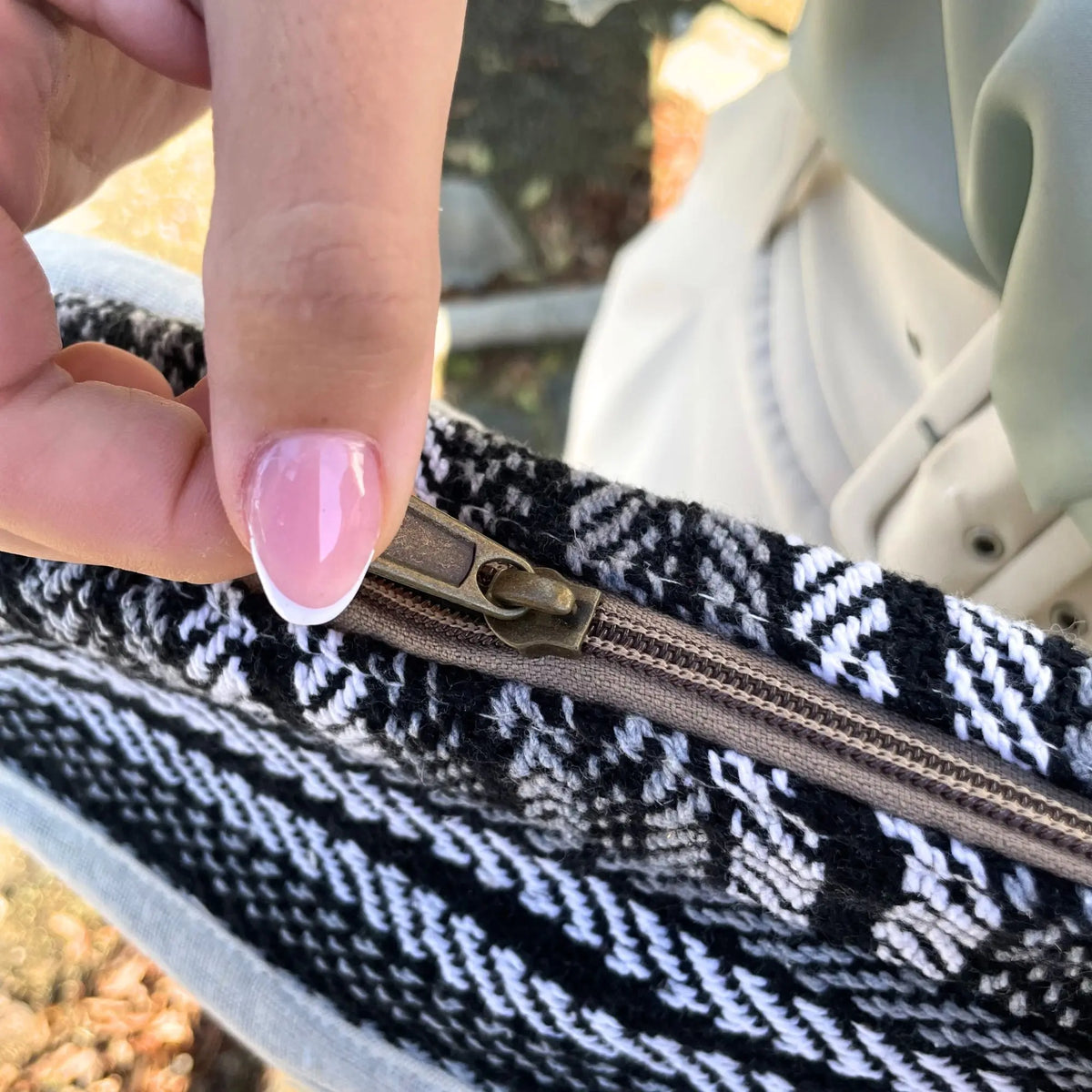 Mano abriendo cremallera de bolso textil tejido con estampado geométrico en blanco, negro y gris.