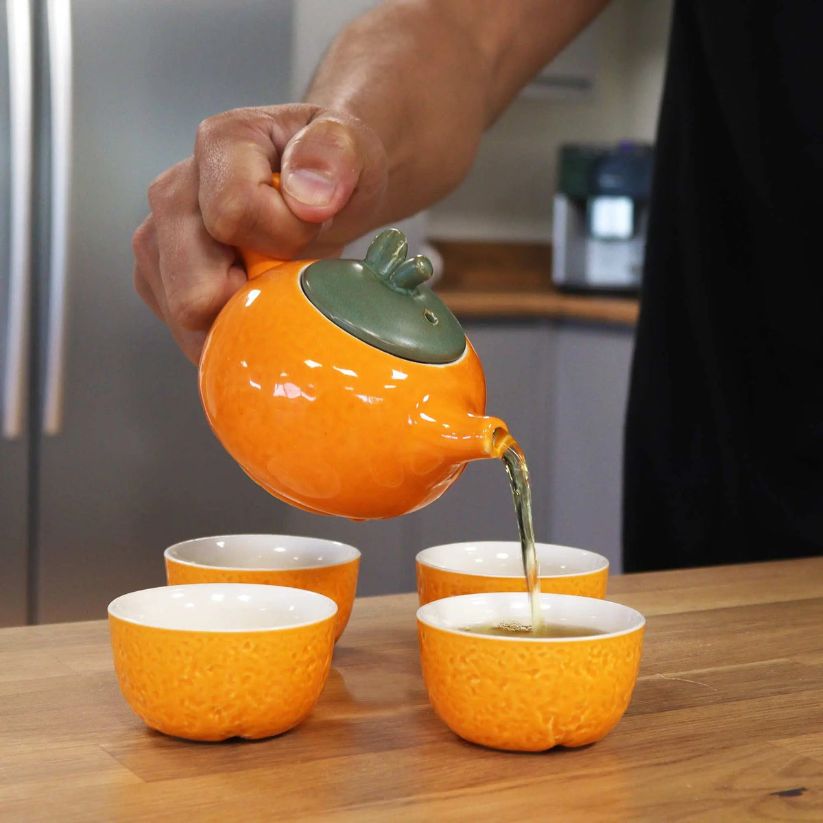 Tetera y tazas de cerámica naranja con textura, vertiendo té en una mesa de madera. Diseño elegante y moderno.
