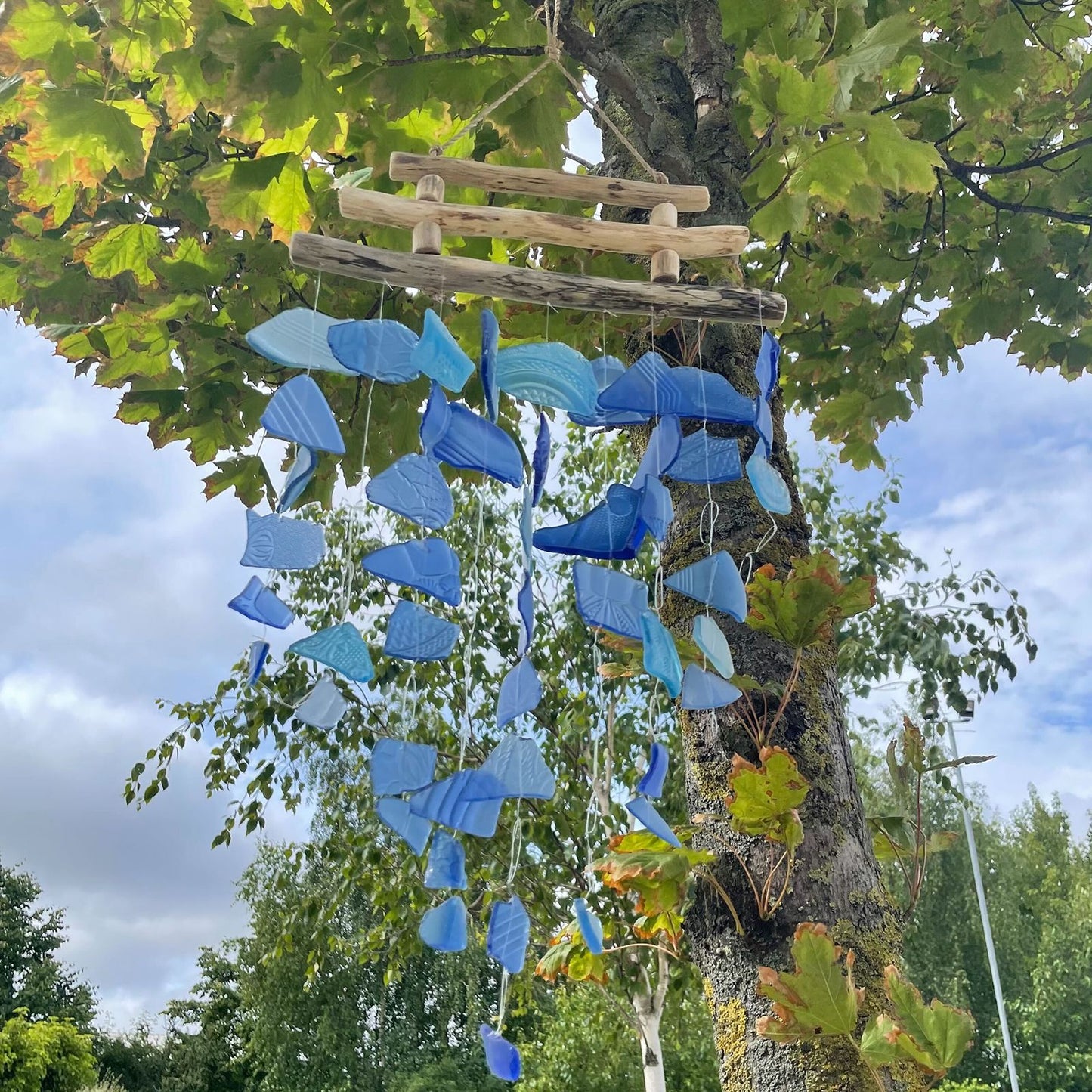 Carrillón de piezas de vidrio reciclado en tonos azules, suspendido de ramas de madera natural en un árbol.