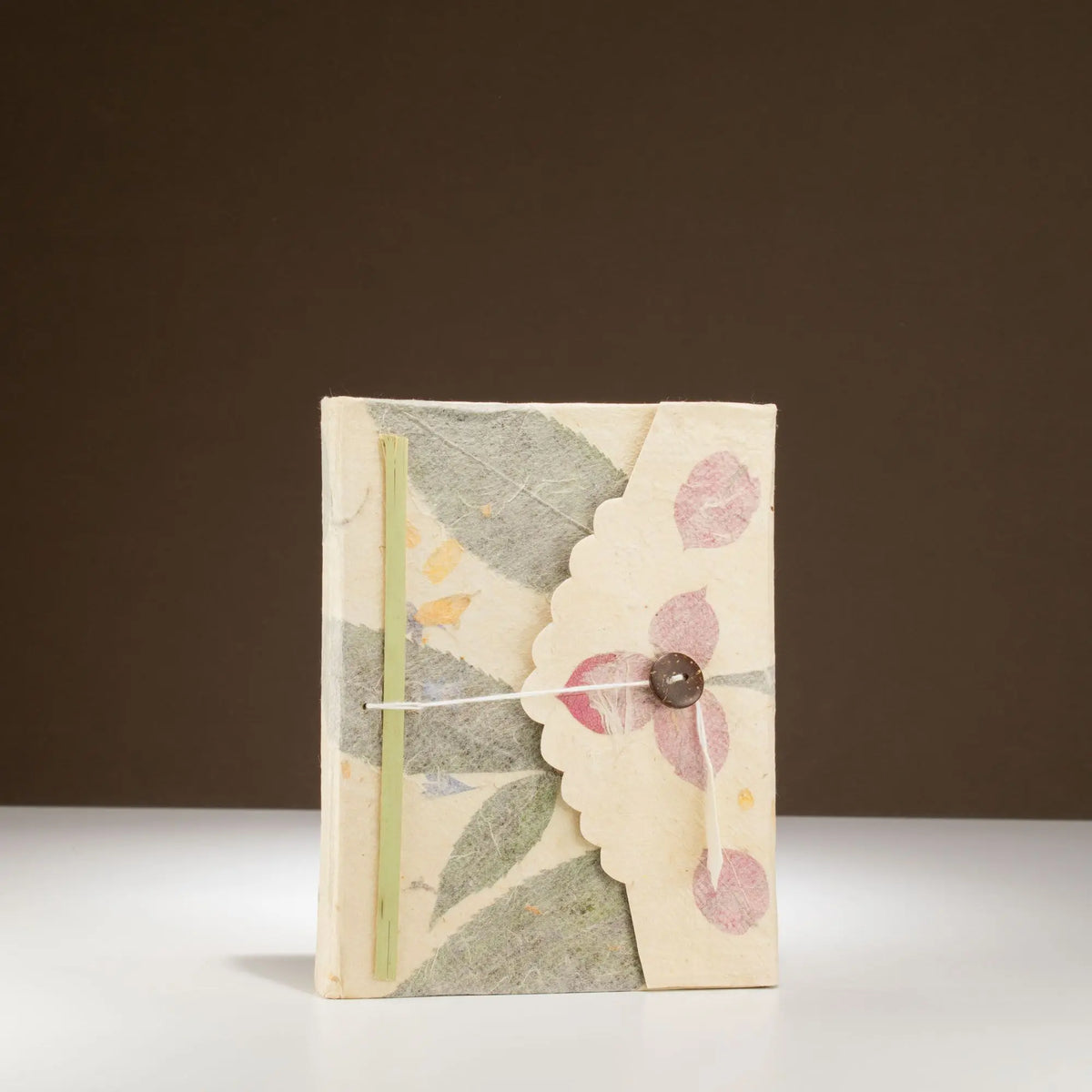 Cuaderno artesanal de papel reciclado con flores rosadas, tapa en tonos crema y cinta de cierre con botón.