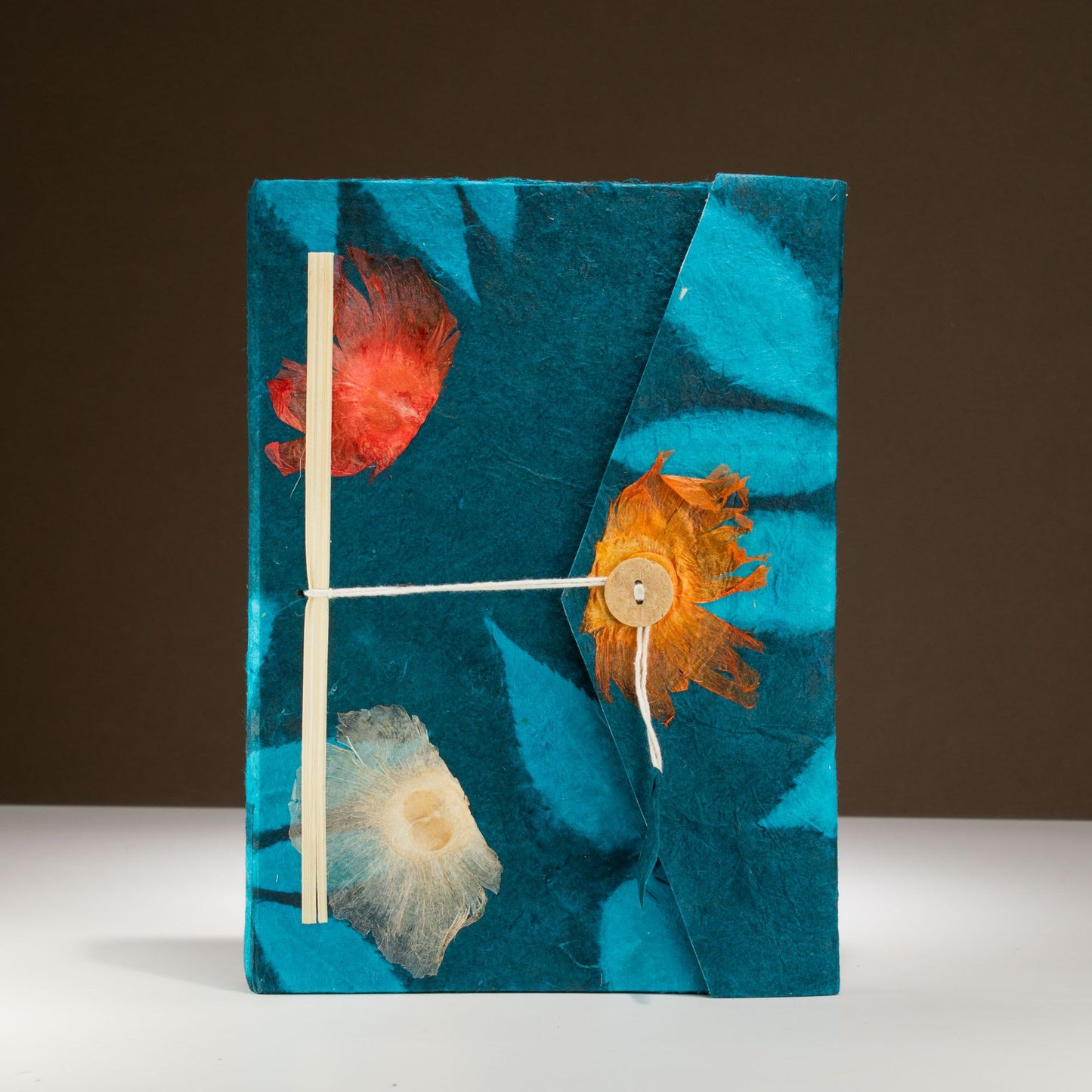 Cuaderno artesanal azul con flores secas en tonos naranja y blanco, encuadernación sencilla con cuerda de yute.
