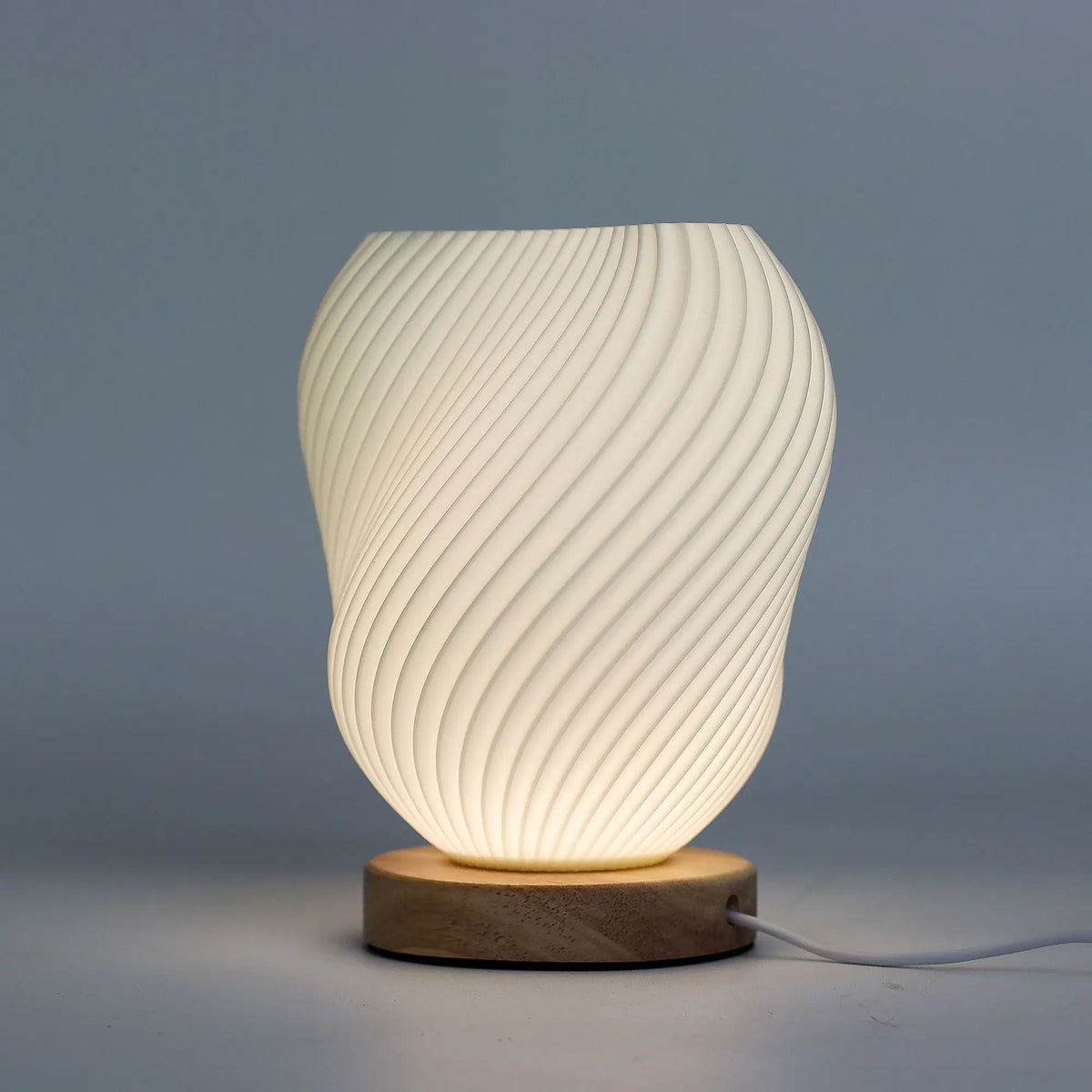 Lámpara blanca de sobremesa con pantalla en forma de concha espiralada sobre una base redonda de madera clara.