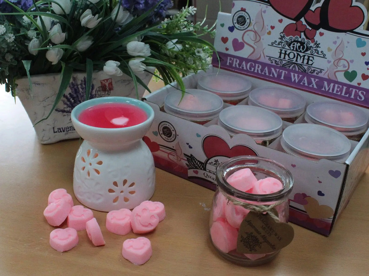 Quemador de cerámica con cera rosa, frasco de vidrio con corazones de cera y caja de exhibición con flores al fondo.