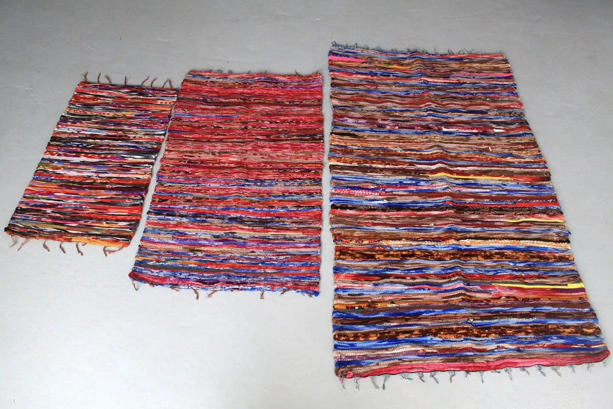 Tres alfombras rectangulares de trapo tejidas a mano con rayas horizontales multicolores y flecos en los extremos.
