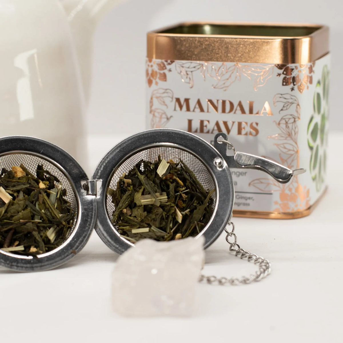 Infusor metálico abierto con té verde, jengibre y limón junto a una lata cuadrada blanca con detalles en cobre.