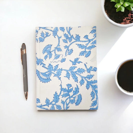 Cuaderno de tela con estampado floral azul sobre fondo crema, junto a un bolígrafo y una taza de café.