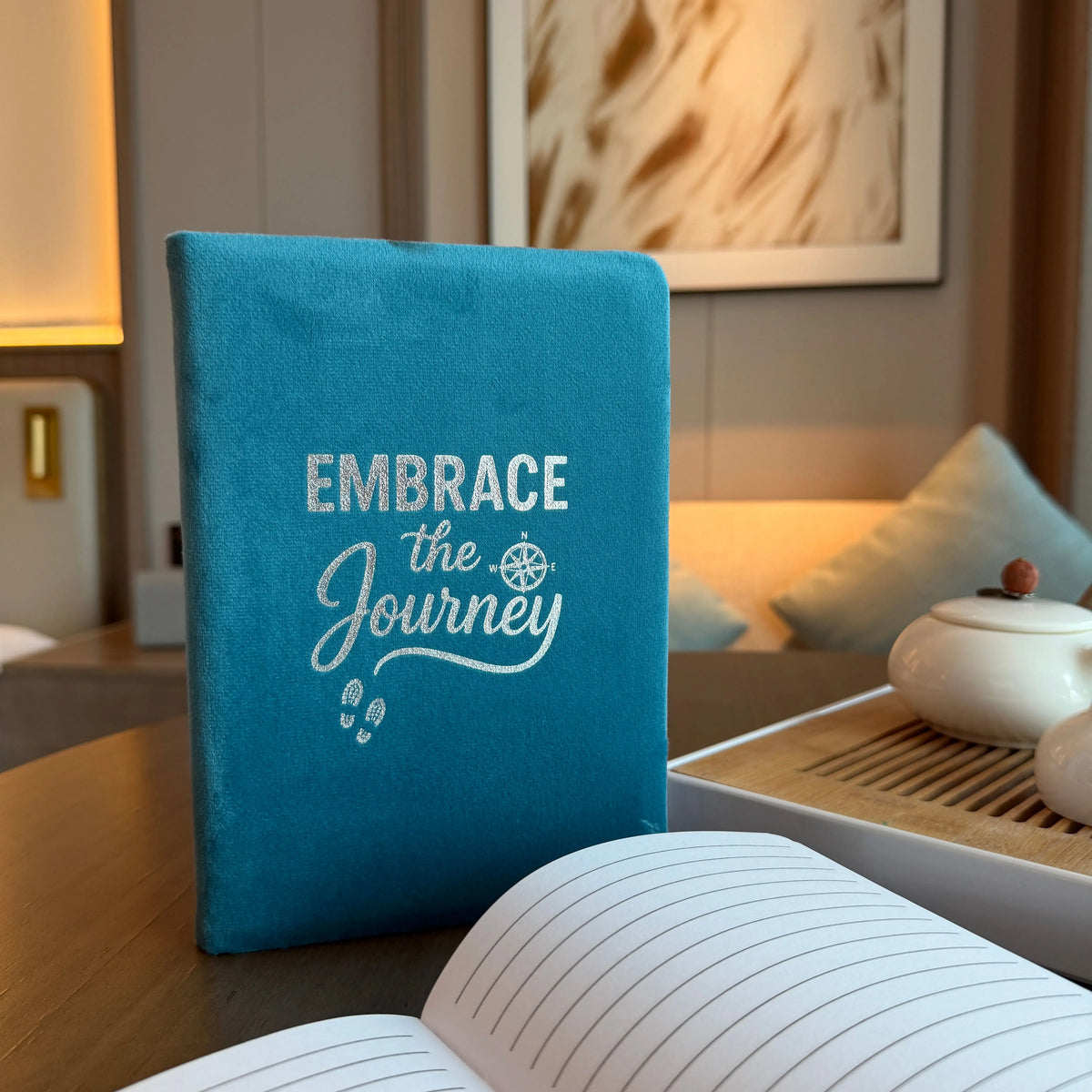Cuaderno de terciopelo azul claro con letras plateadas "Embrace the Journey" sobre una mesa, junto a un té.