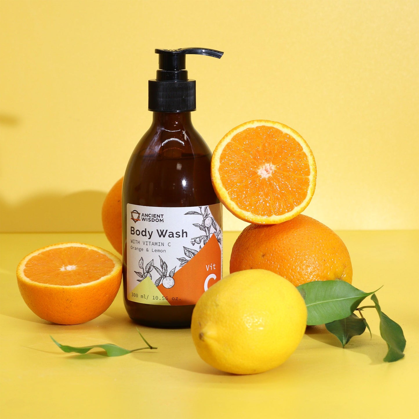Jabón Ancient Wisdom Vitamina C en frasco oscuro con dispensador, rodeado de naranjas y un limón sobre fondo amarillo.