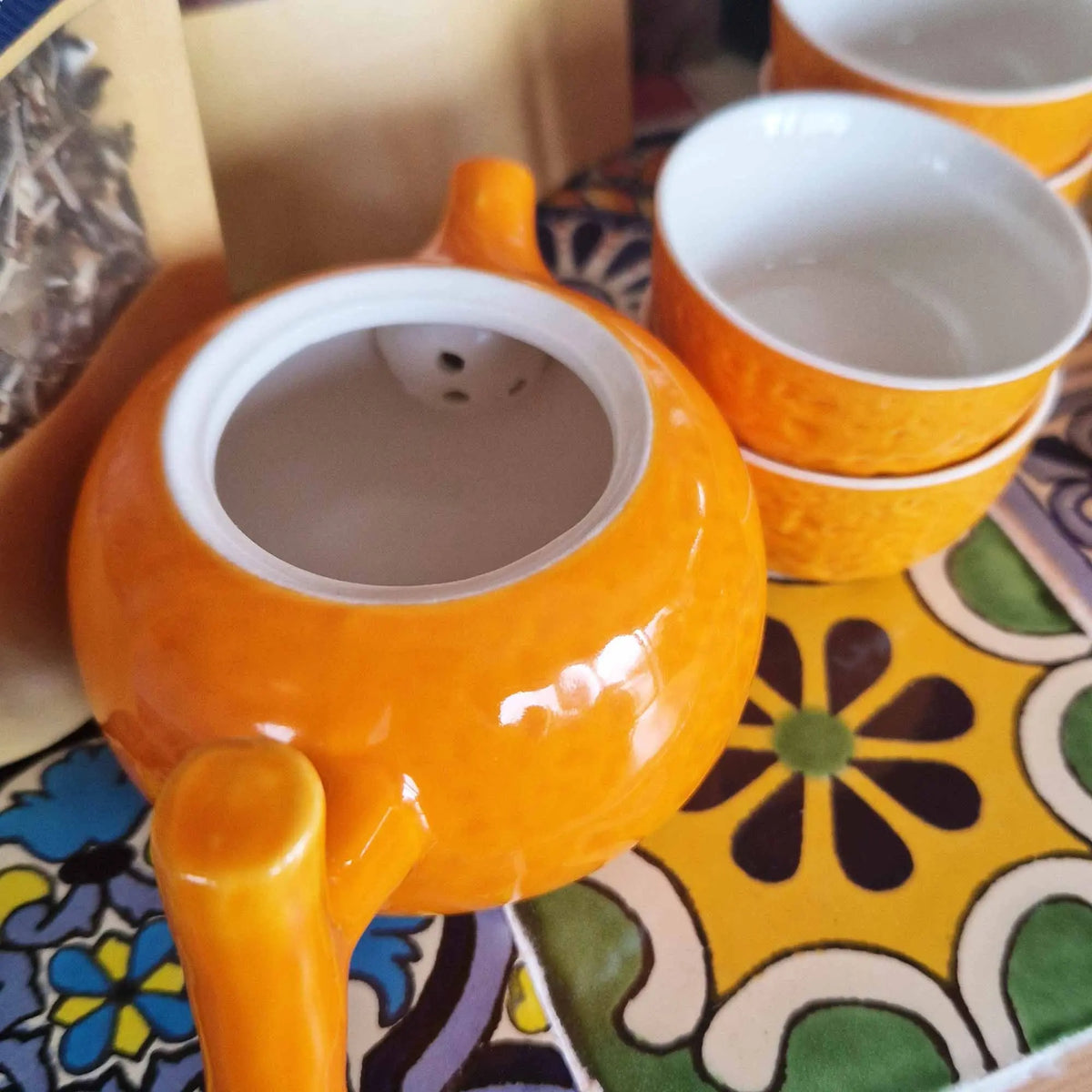Tetera de cerámica naranja brillante con filtro integrado y dos tazas a juego, sobre un fondo colorido.