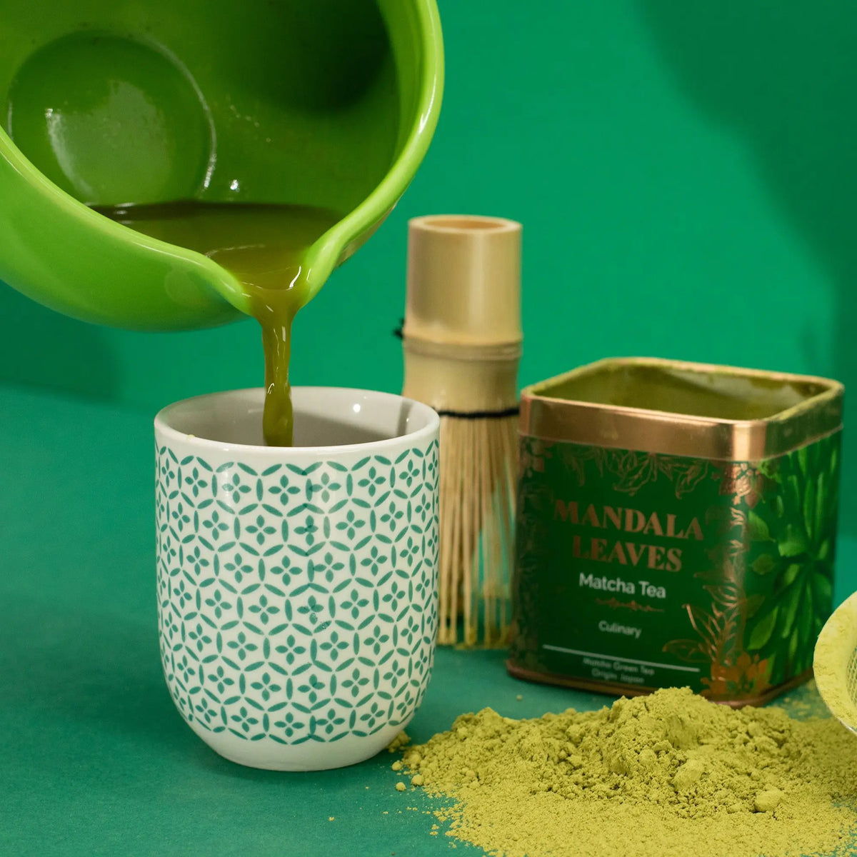 Se sirve matcha en una taza estampada junto a una lata verde de té, un batidor de bambú y polvo de té sobre fondo verde.
