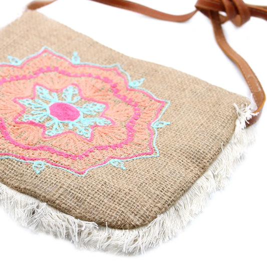 Bolso de yute con bordado tipo mandala en tonos rosa y azul, detalles de flecos blancos y correa de cuero marrón.