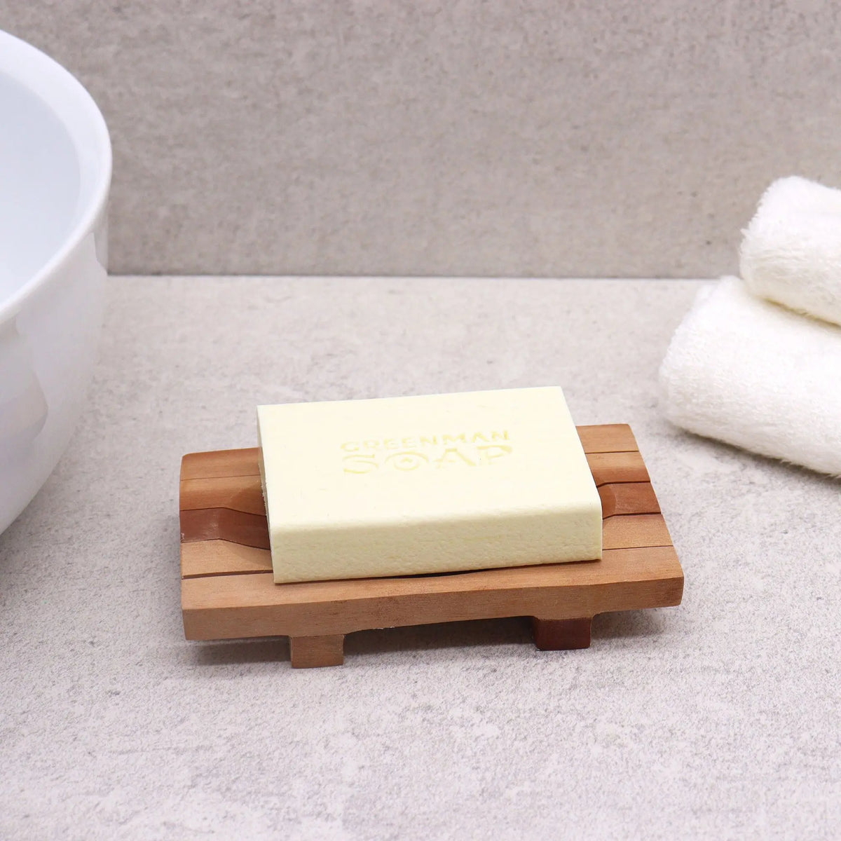 Jabonera de madera de caoba con rejilla drenante y pastilla de jabón sobre superficie clara de baño.