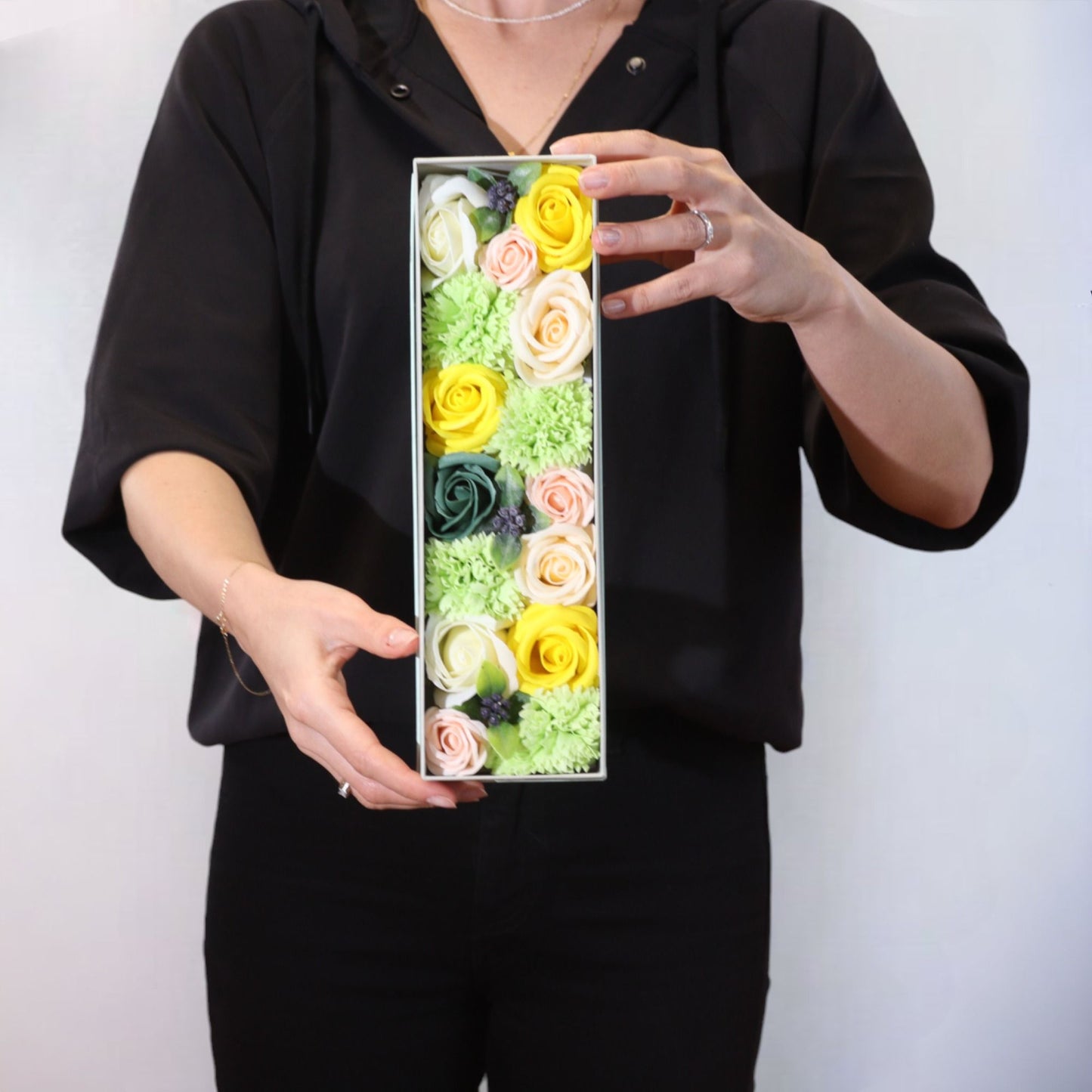 Caja rectangular con flores de jabón en tonos amarillos, verdes y rosas, sostenida por una persona vestida de negro.
