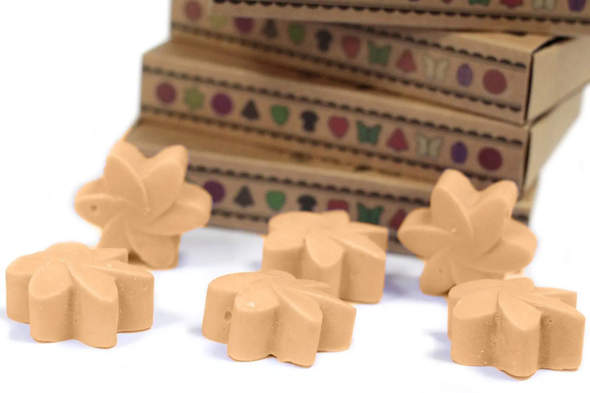 Seis ceras de soja beige con forma de flor frente a cajas de cartón apiladas con motivos geométricos coloridos.