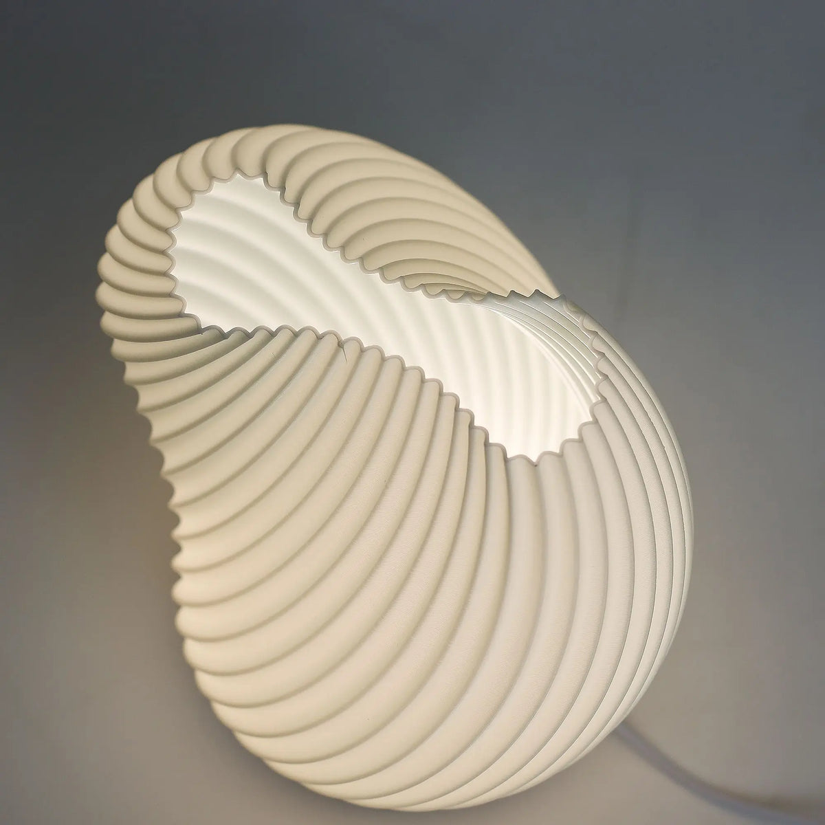 Lámpara blanca de mesa con forma de concha marina, diseño en espiral estriado y luz interior suave y difusa.