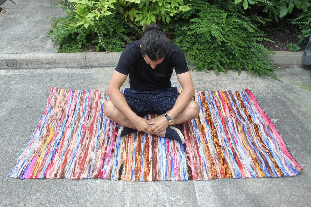 Persona sentada sobre una alfombra de trapo rectangular, multicolor y de tejido artesanal indio, de 105x185 cm.