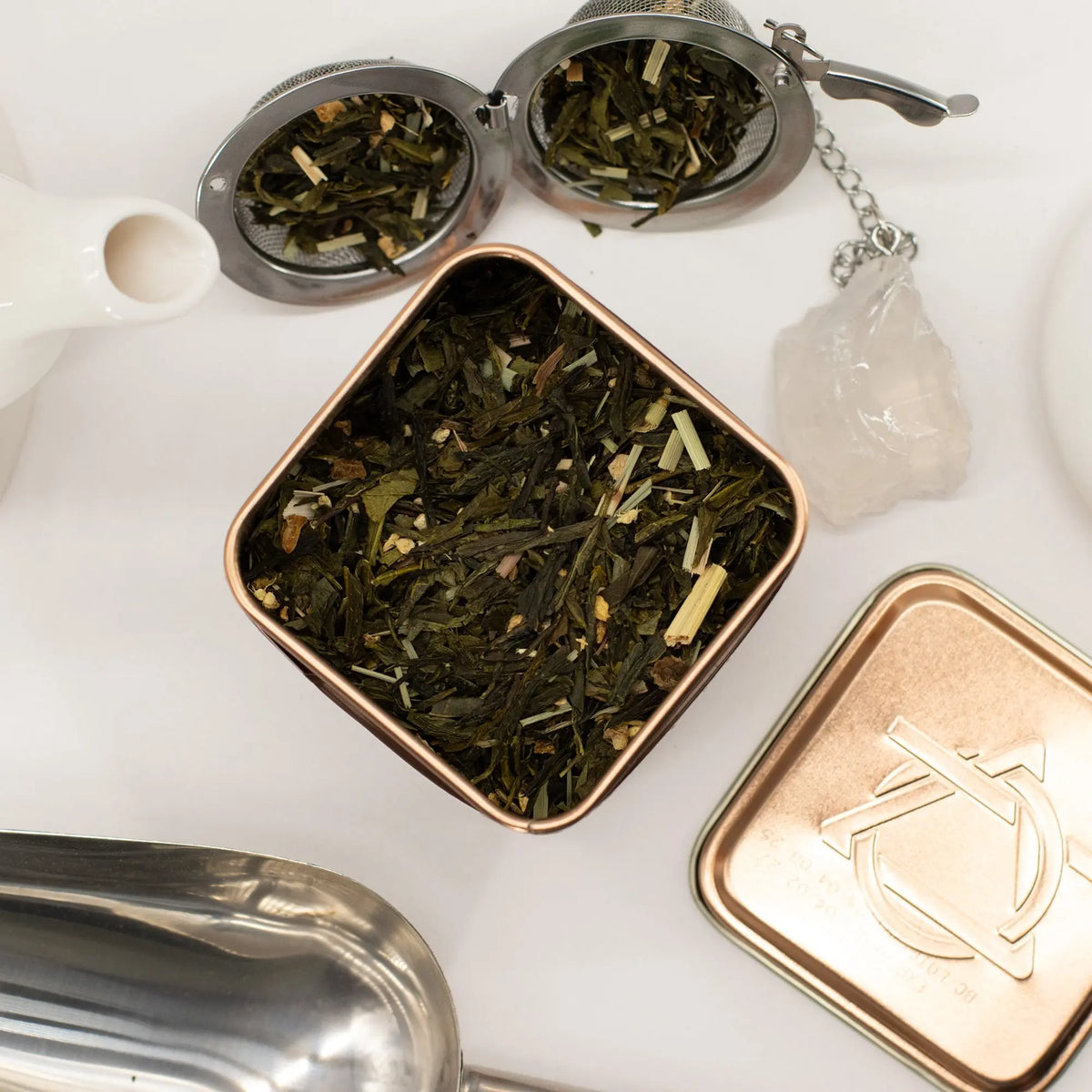 Lata cuadrada de color cobre abierta con té verde, jengibre y limón, acompañada de infusores y una cuchara metálica.