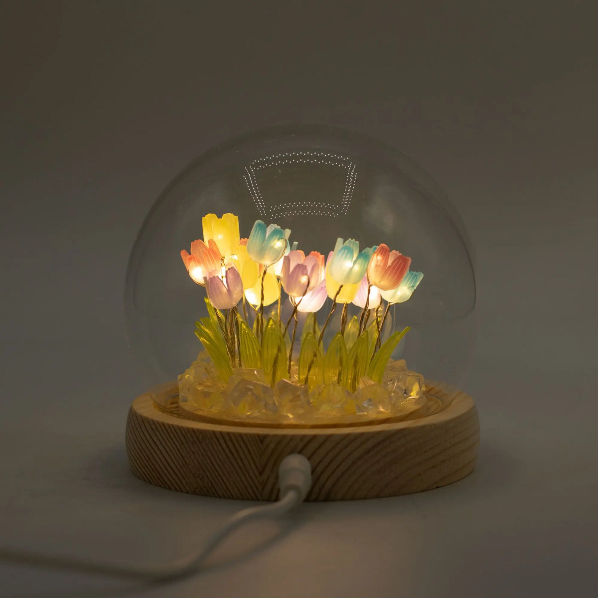 Domo de cristal con tulipanes LED multicolores sobre base de madera clara, iluminado mediante conexión USB.