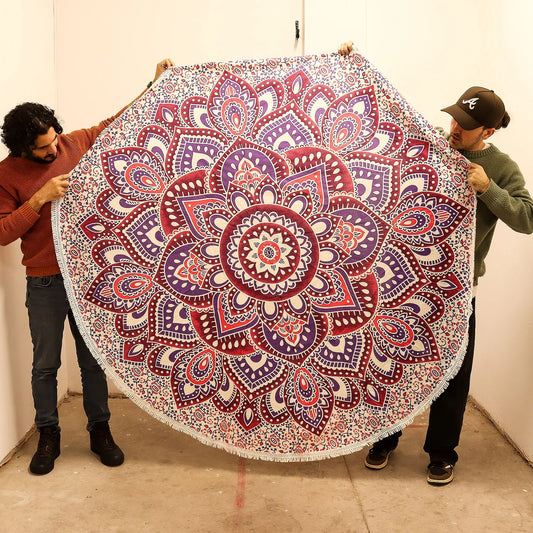 Tapiz redondo de algodón (180 cm) con diseño de mandala en tonos rosa y morado, sostenido por dos personas.