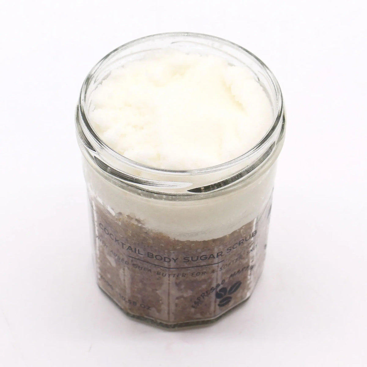Exfoliante de azúcar en frasco de vidrio; capa superior blanca cremosa y base granular oscura tipo café. Fondo blanco.