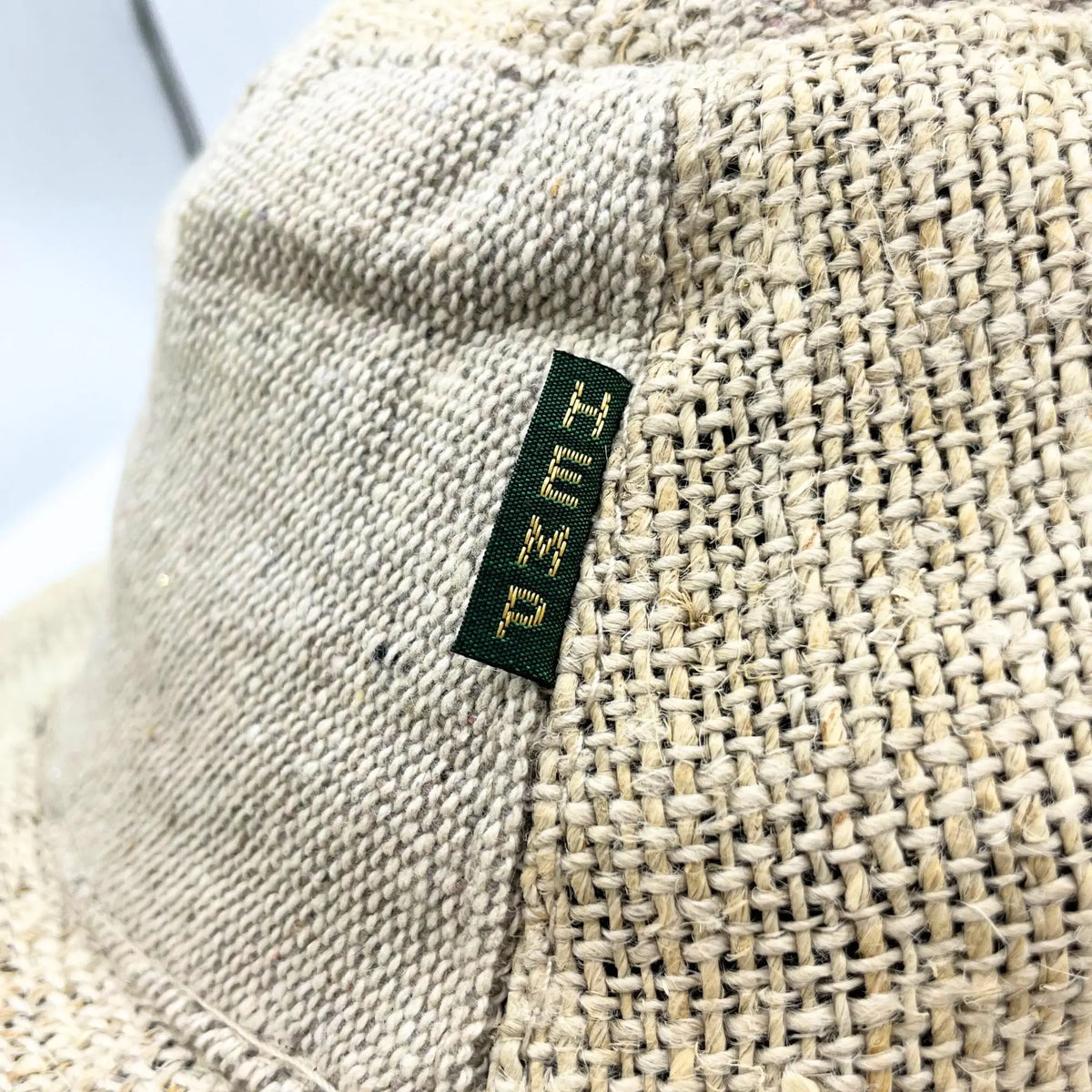Detalle de sombrero color natural con textura de cáñamo tejido y una etiqueta textil verde con letras bordadas.